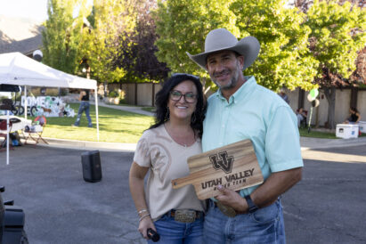 A smiling couple stands outdoors, one holding a wooden sign that reads “Utah Valley Rodeo Team,” with a festive event backdrop featuring tents and trees.